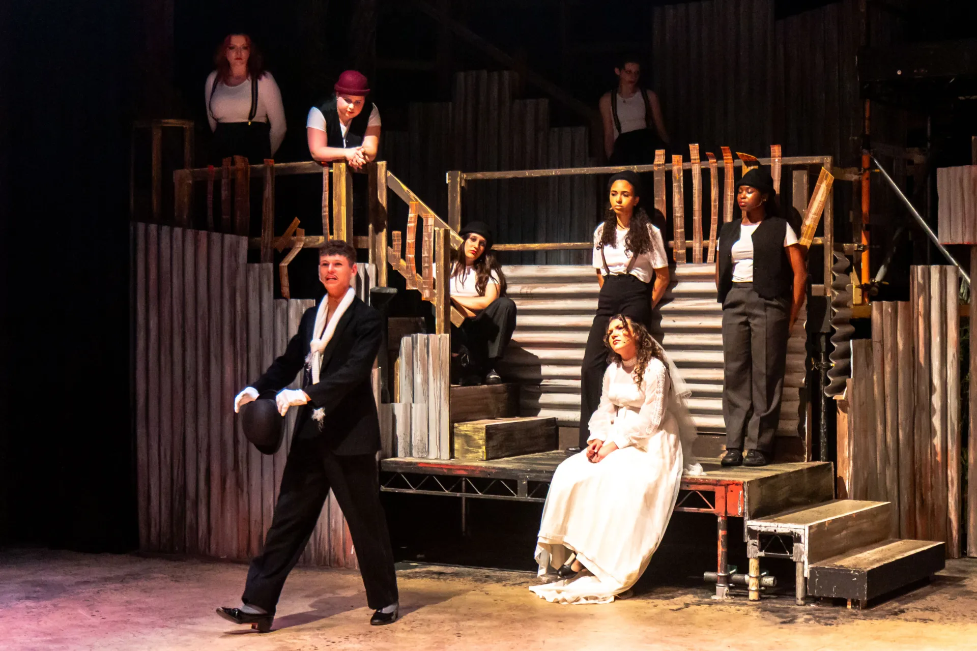 An actor in a tuxedo and white scarf walks across the stage while a woman in a wedding dress and a group of performers in vests and suspenders look on from a multi-level industrial set.