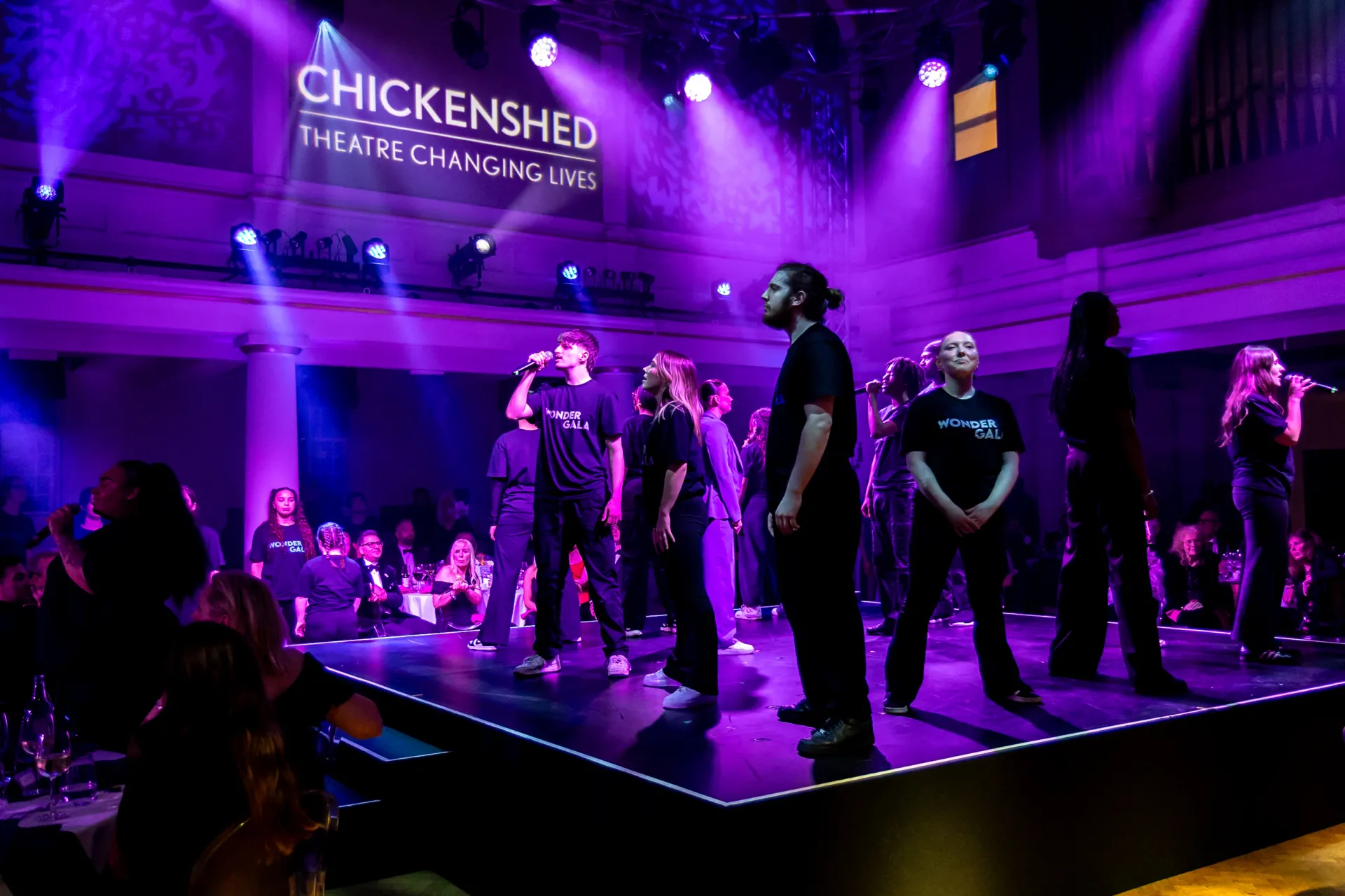 A group of performers wearing black "WONDER GALA" shirts sing on a stage lit by purple spotlights, with the "CHICKENSHED" logo projected in the background.