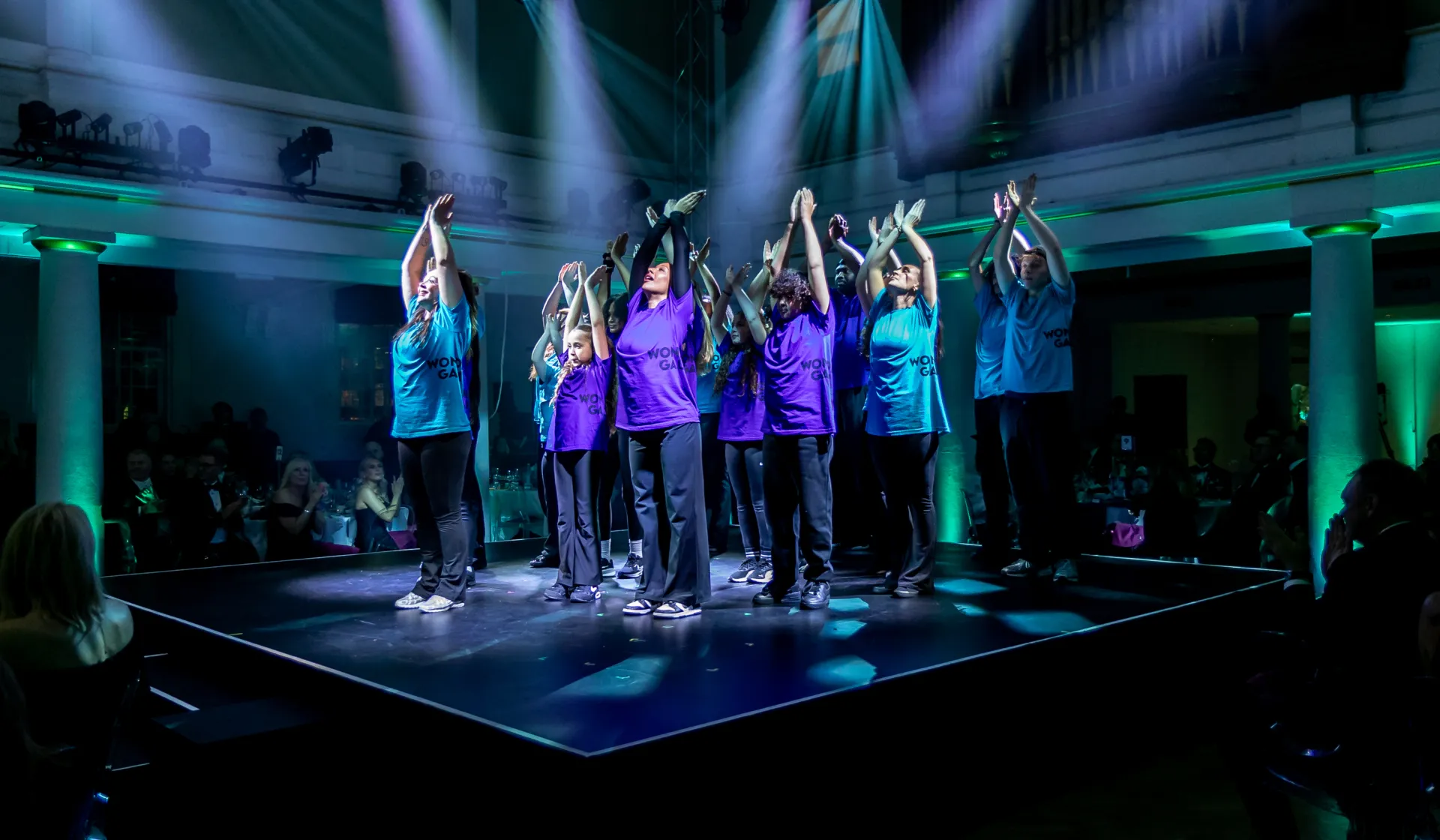 Group of performers with arms up on stage at the Wonder Gala, purples and blues, joyful and hopeful