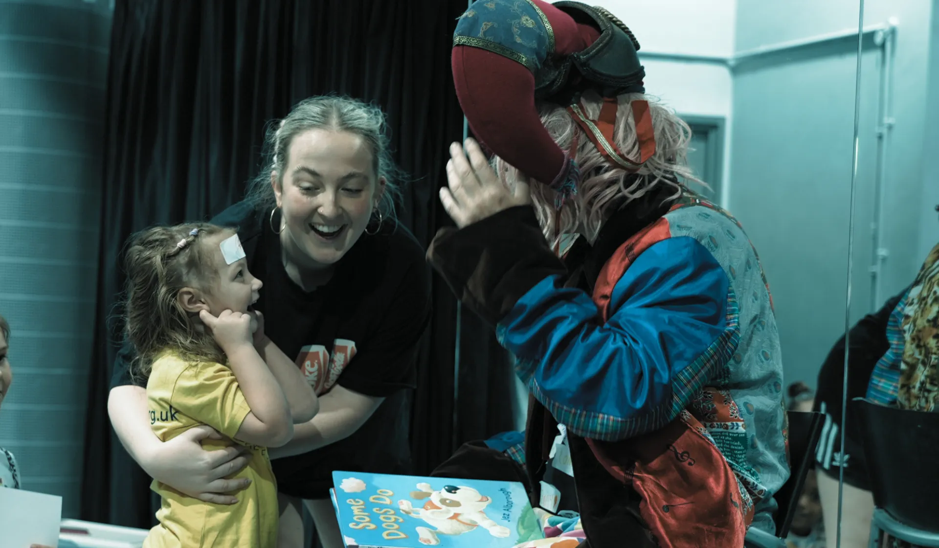 A man in a jester hat talking to a young girl who is smiling and being held by a woman.