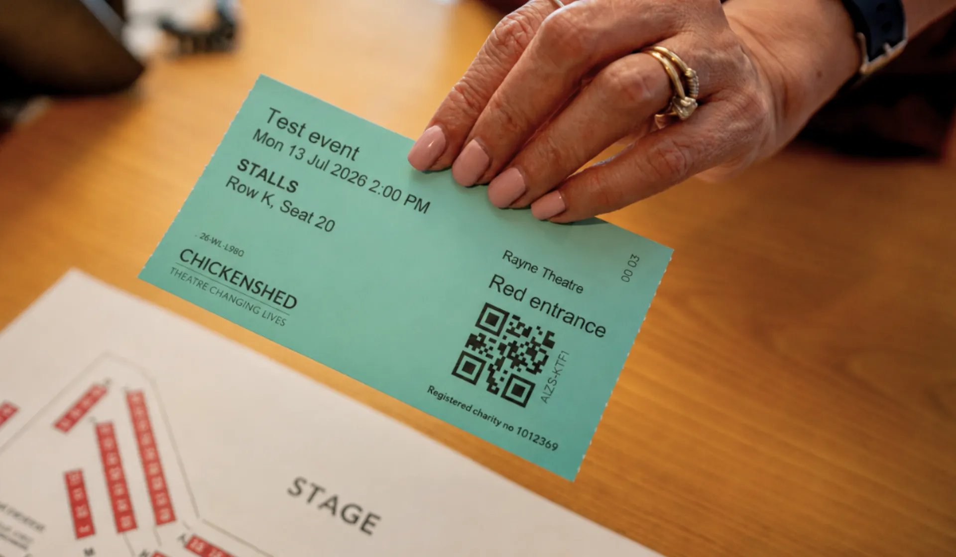 A hand holds a teal Chickenshed theatre ticket above a seating plan on a wooden desk. The ticket shows a test event at the Rayne Theatre, with details for Stalls, Row K, Seat 20, and a QR code.