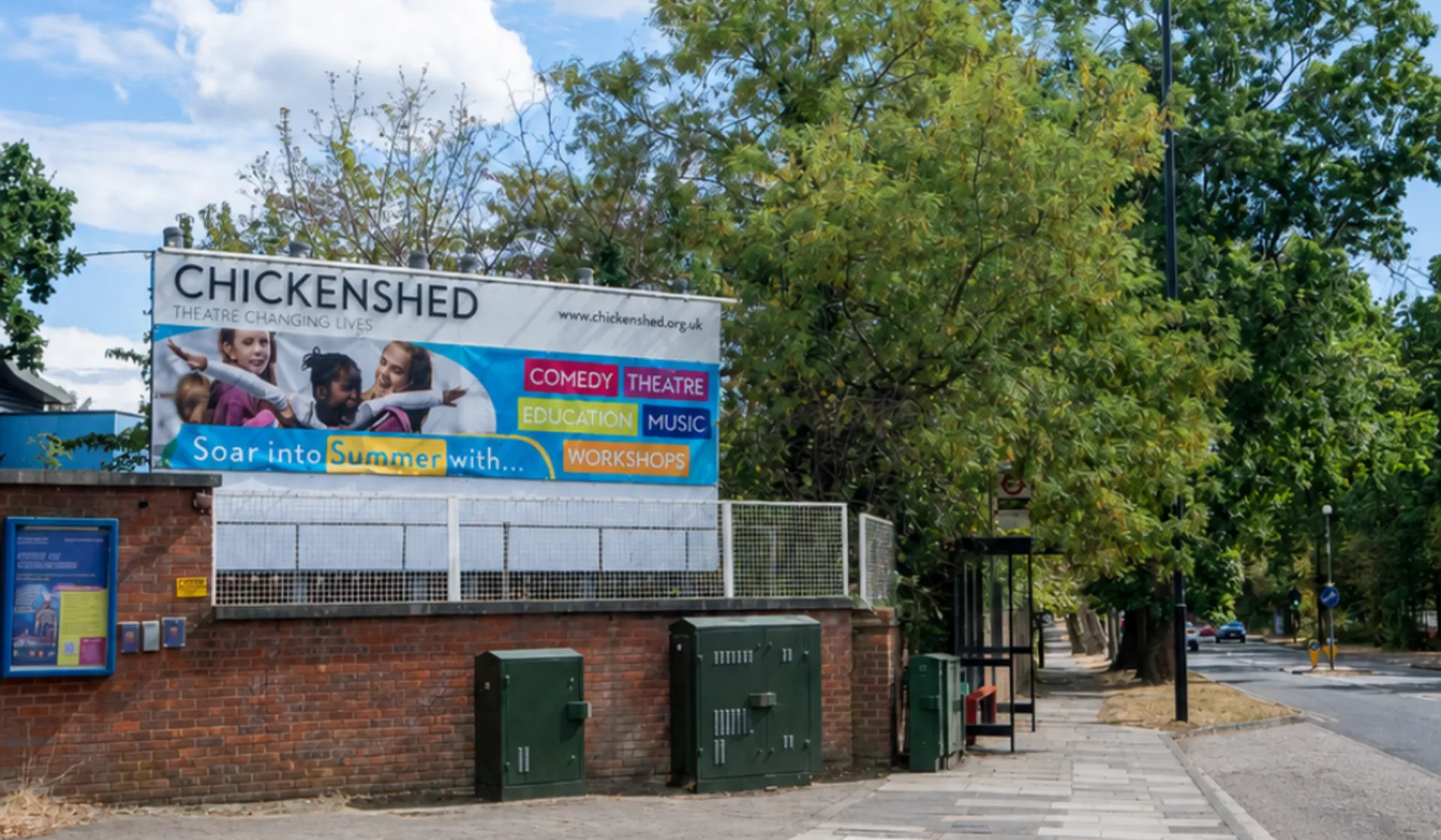 Chickenshed Theatre’s colourful outdoor banner on a brick wall beside a tree-lined street and pavement, beneath a bright blue sky with white clouds.