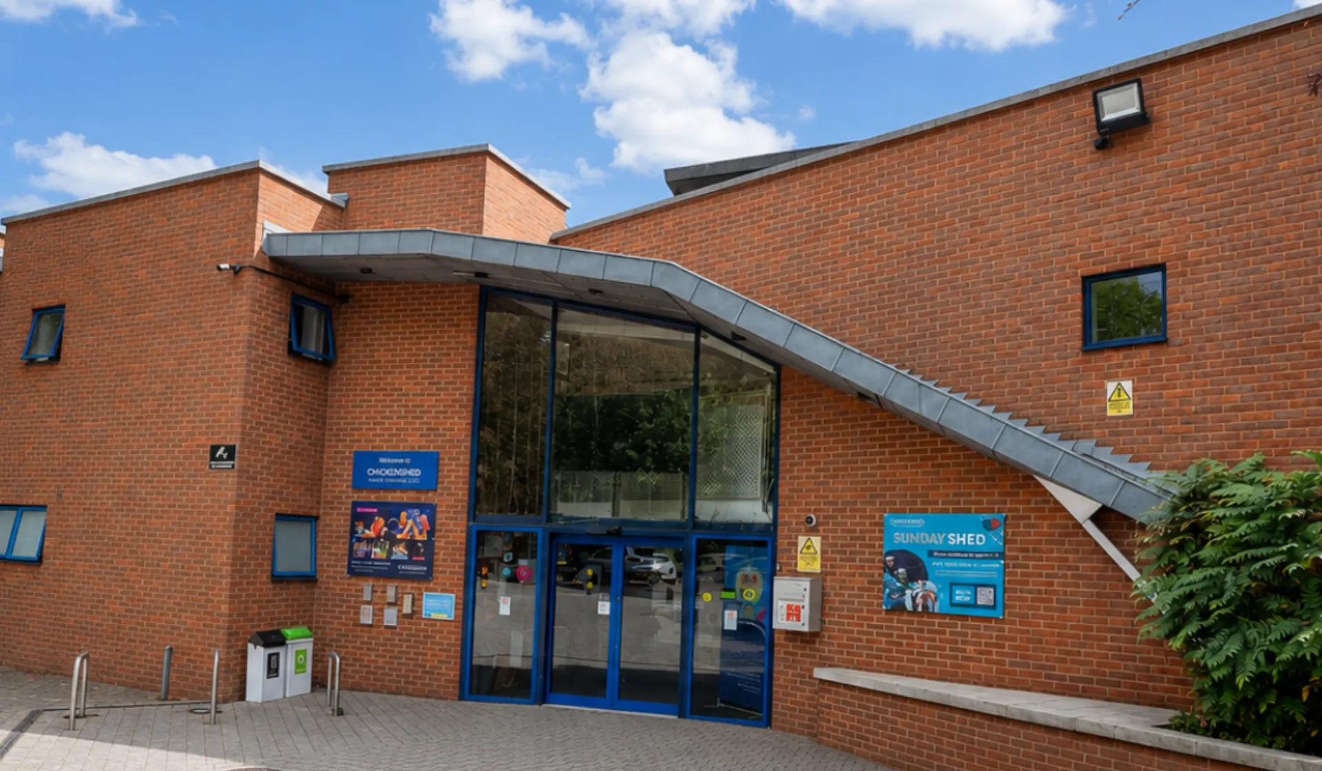 Chickenshed Theatre’s red-brick building entrance on a sunny day, surrounded by trees and greenery, with blue-trimmed windows, a glass entrance and colourful event signage.