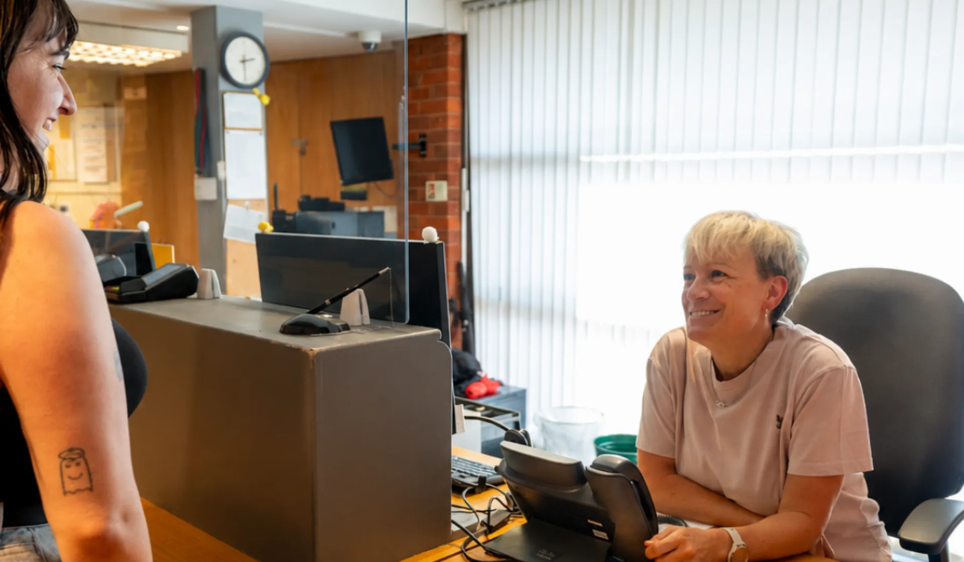Two people chatting at Chickenshed Theatre’s reception desk, with one person seated behind a glass partition and a seating plan visible on the desk.