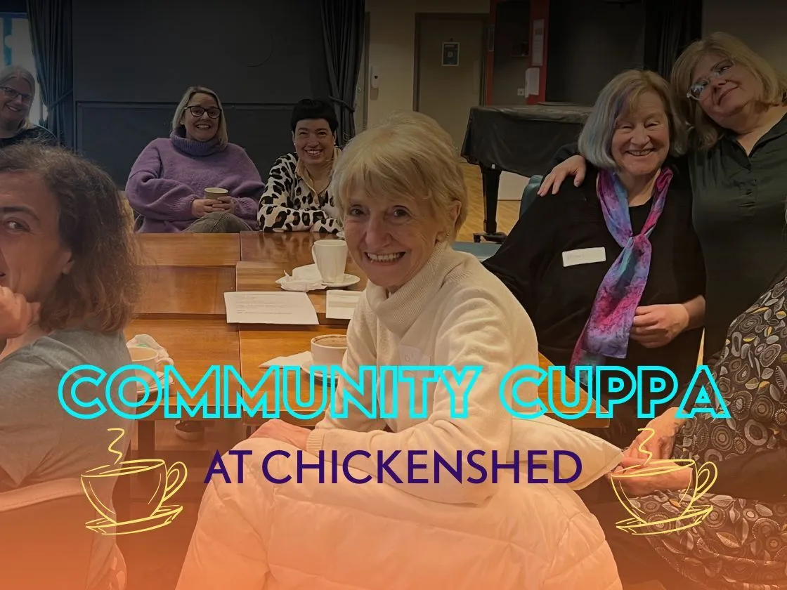 A diverse group of people sitting together in Chickenshed’s café, smiling and chatting around a table during a warm, inclusive Community Cuppa gathering.