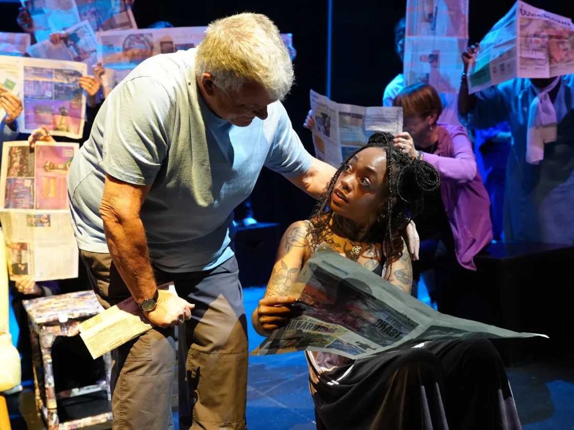 Woman in chair reading newspaper, looking at a man leaning in from behind her