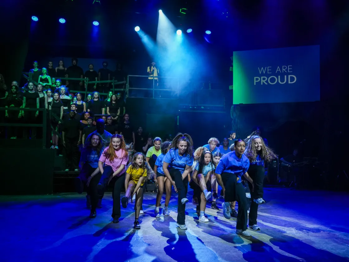 group of performers mid dance on stage with chorus of singers lifted onto back of stage