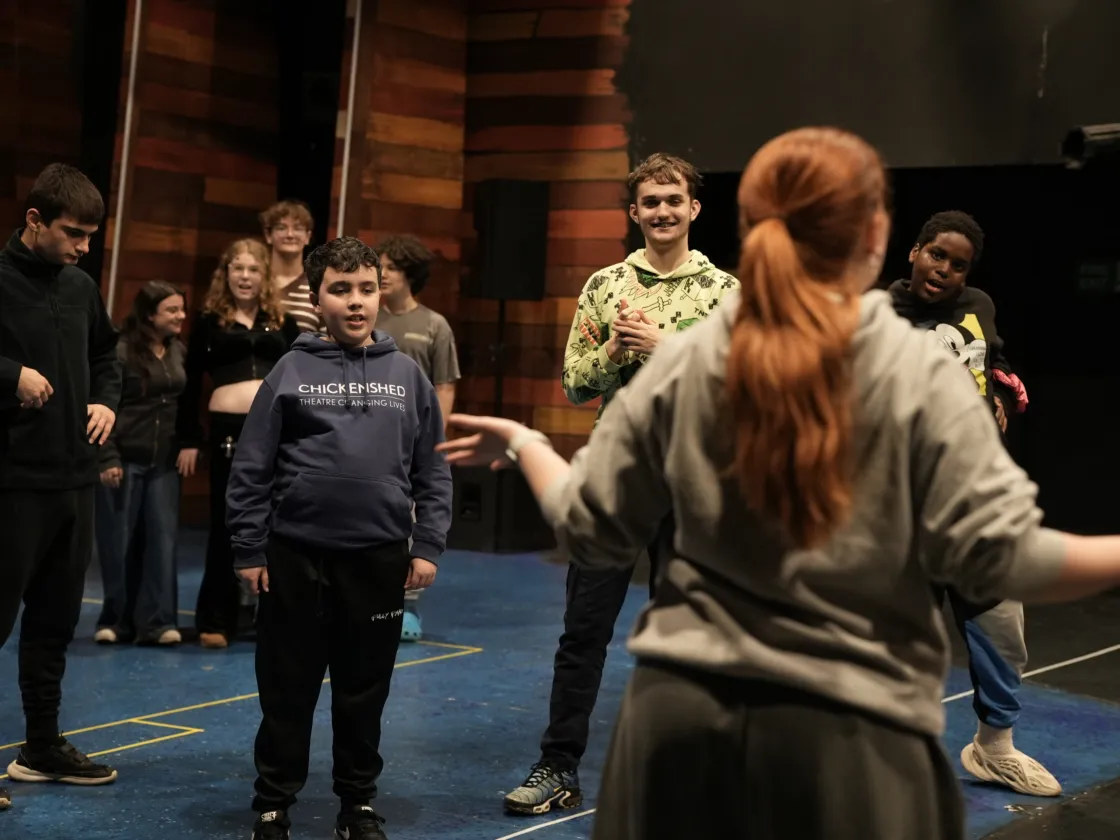 A teacher at the front of the room with bright orange hair leading the rest of the class of children in a dance
