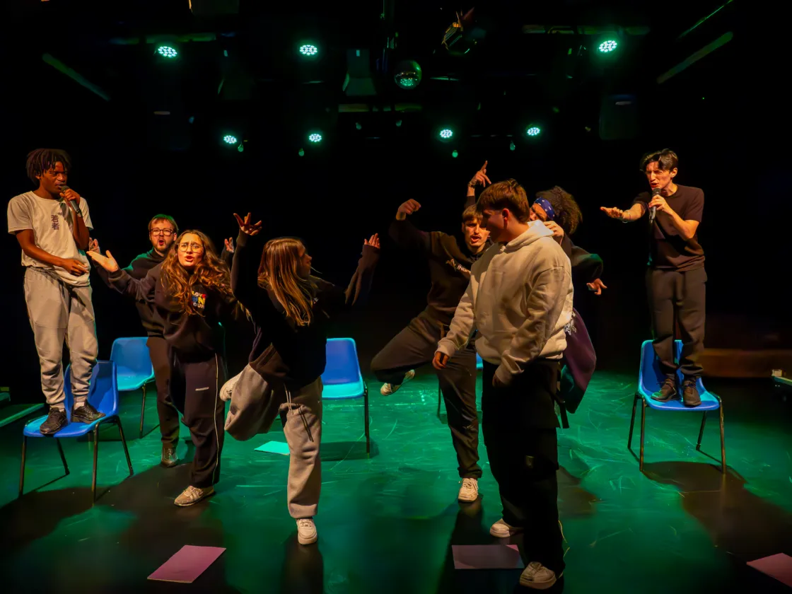 A group of young performers in casual streetwear act out a spirited scene under green stage lights, with two individuals standing on chairs and holding microphones.