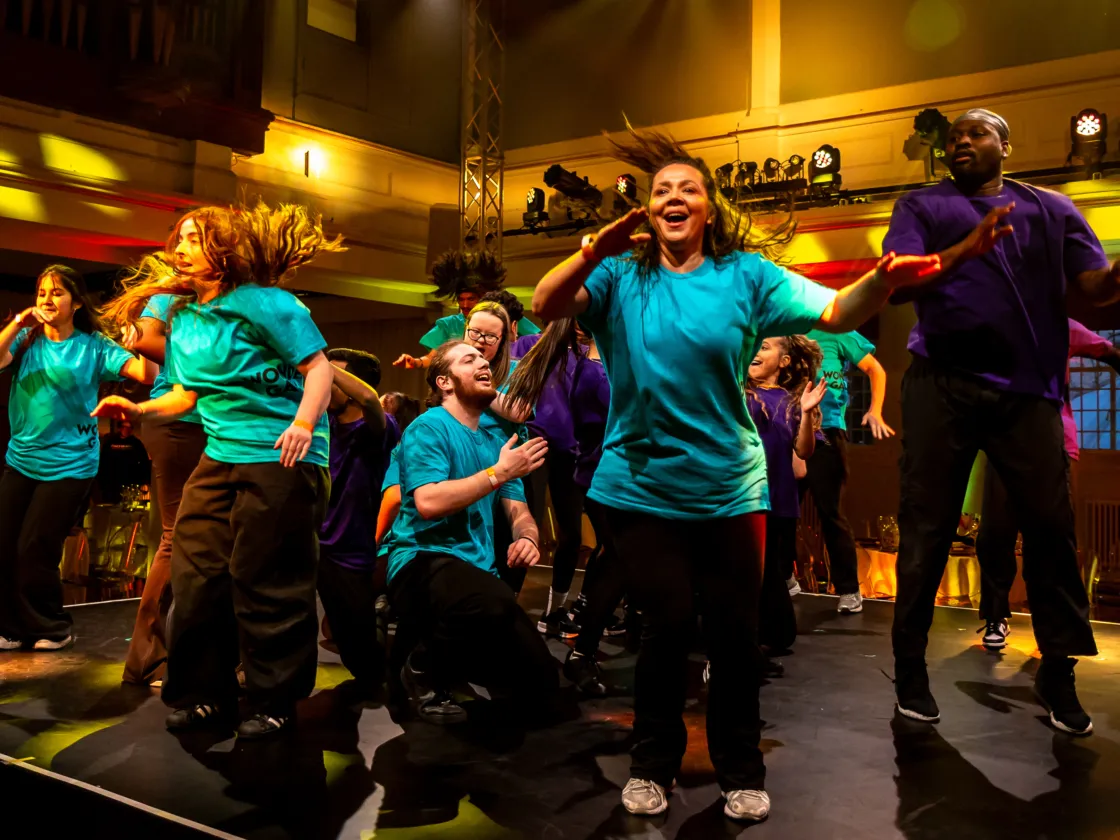 A joyful ensemble of performers in bright teal and purple t-shirts dance energetically on a stage during the "Wonder Gala," illuminated by warm yellow theatrical lighting.