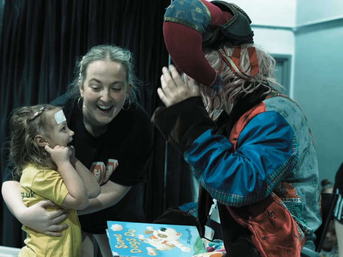 A man in a jester hat talking to a young girl who is smiling and being held by a woman.
