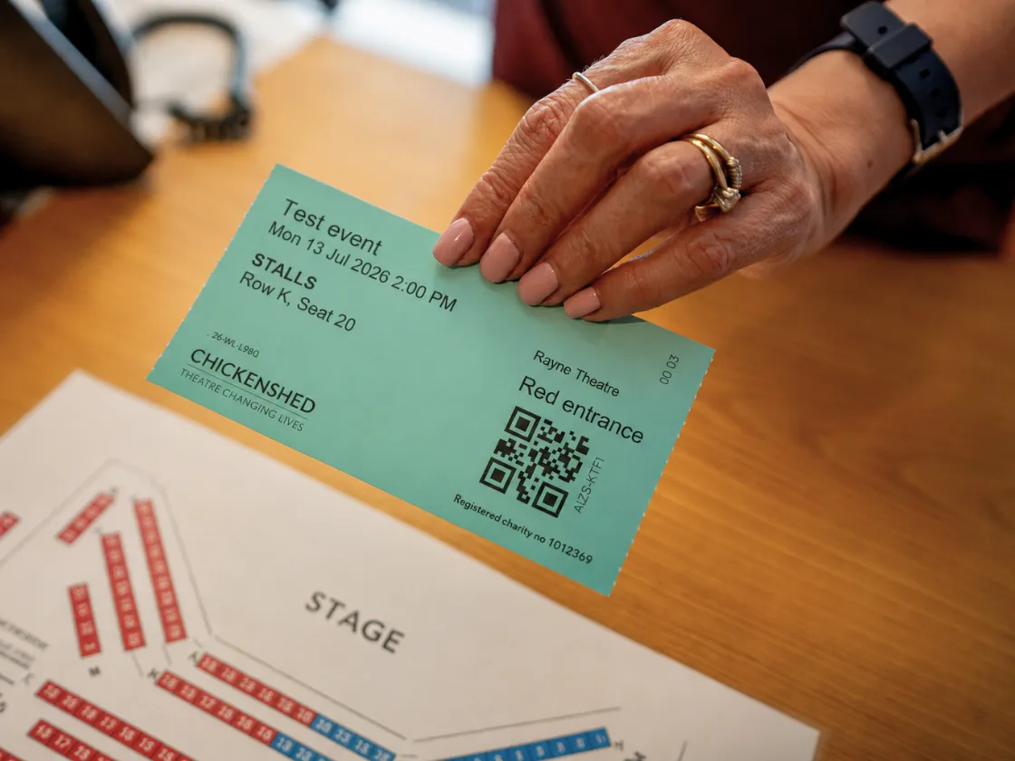 A hand holds a teal Chickenshed theatre ticket above a seating plan on a wooden desk. The ticket shows a test event at the Rayne Theatre, with details for Stalls, Row K, Seat 20, and a QR code.