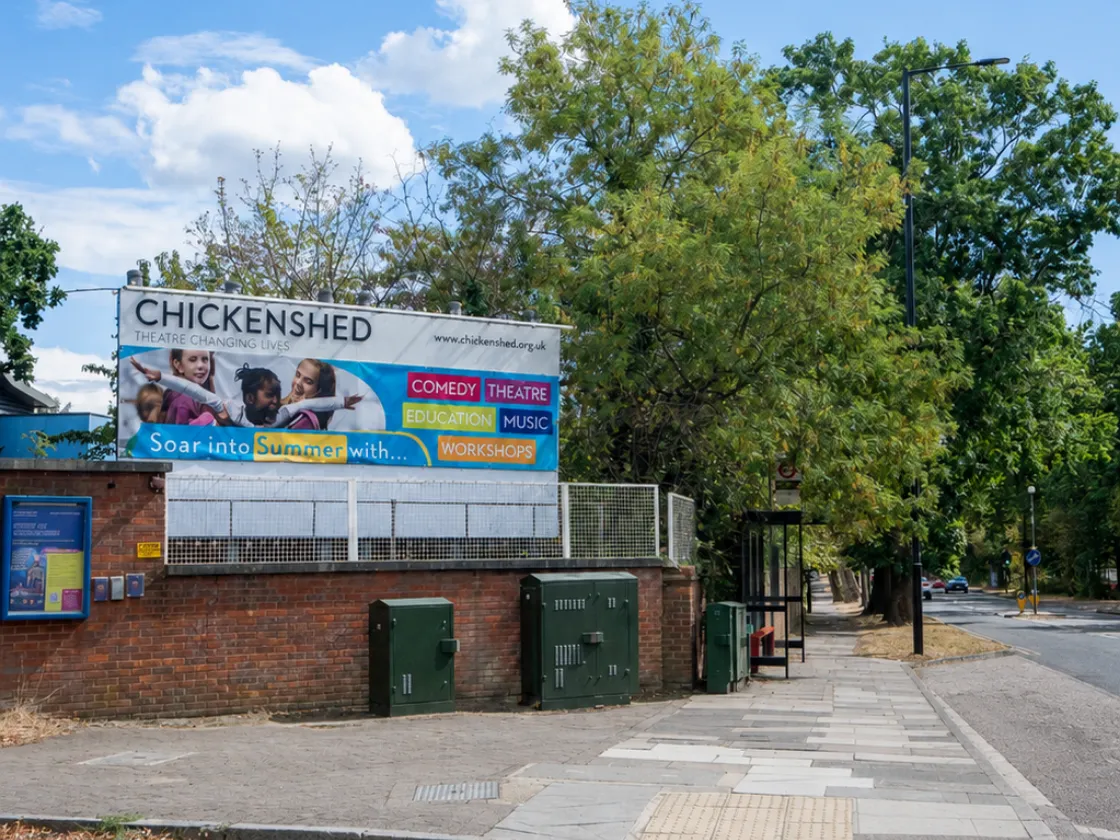 Chickenshed Theatre’s colourful outdoor banner on a brick wall beside a tree-lined street and pavement, beneath a bright blue sky with white clouds.