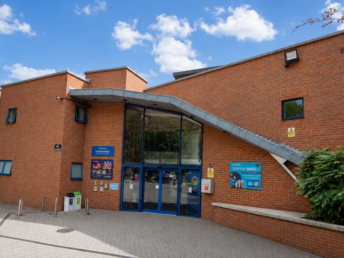 Chickenshed Theatre’s red-brick building entrance on a sunny day, surrounded by trees and greenery, with blue-trimmed windows, a glass entrance and colourful event signage.