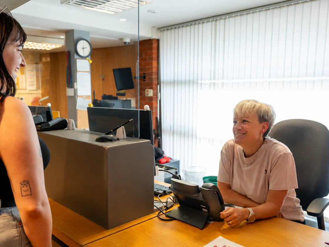 Two people chatting at Chickenshed Theatre’s reception desk, with one person seated behind a glass partition and a seating plan visible on the desk.