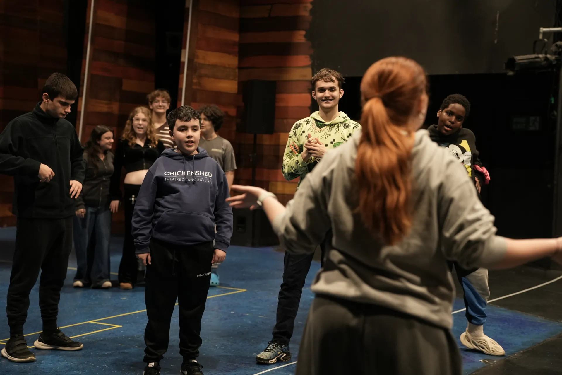 A teacher at the front of the room with bright orange hair leading the rest of the class of children in a dance