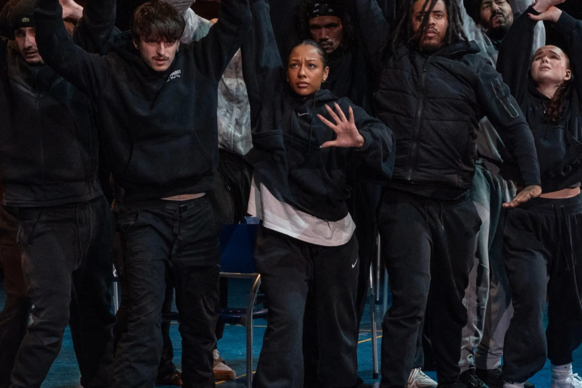 A group of dancers in black hoodies and joggers perform a synchronized routine on a blue stage. They have their arms raised with open palms and intense expressions. The lighting is dramatic, highlighting their powerful movement.
