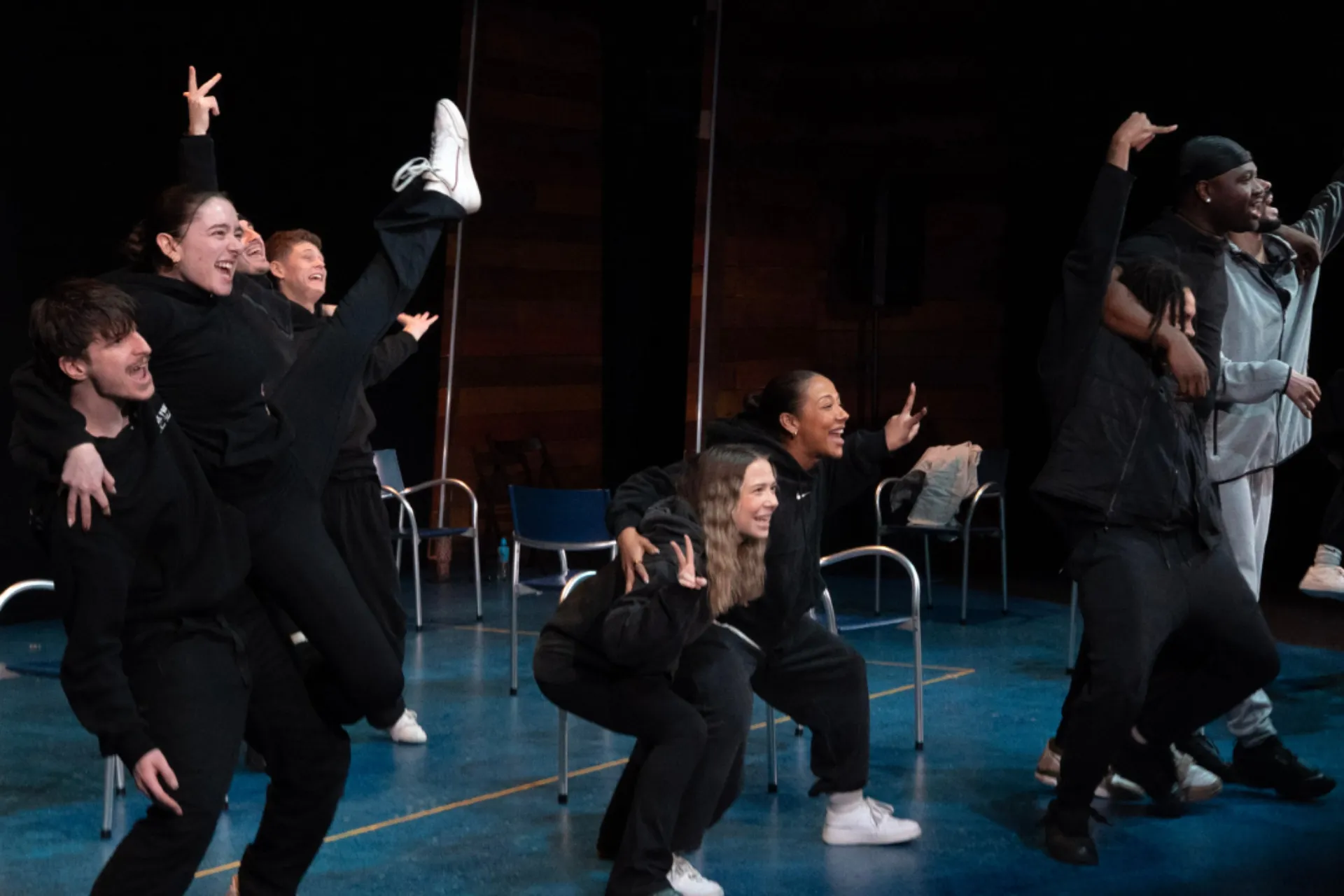 A group of dancers in black streetwear celebrate joyfully on a blue stage. They are laughing and posing together, with some making peace signs and one dancer kicking a leg high into the air. Empty blue chairs are scattered in the background.