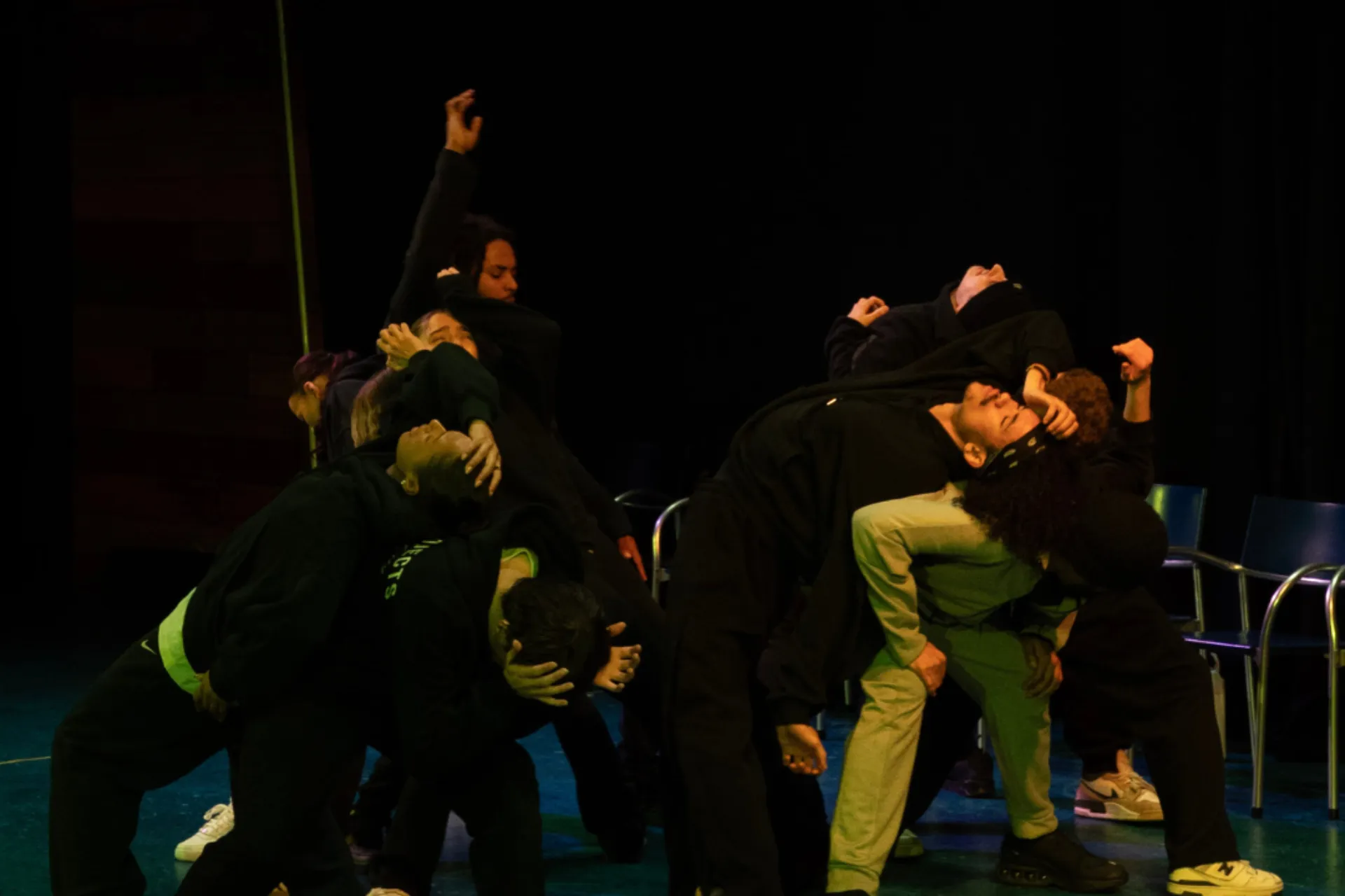 A group of dancers in dark streetwear are huddled together in a dramatic, leaning pose on a blue stage. Their bodies are intertwined, with some holding their heads or reaching out, creating a sense of tension and emotion under low, moody lighting.