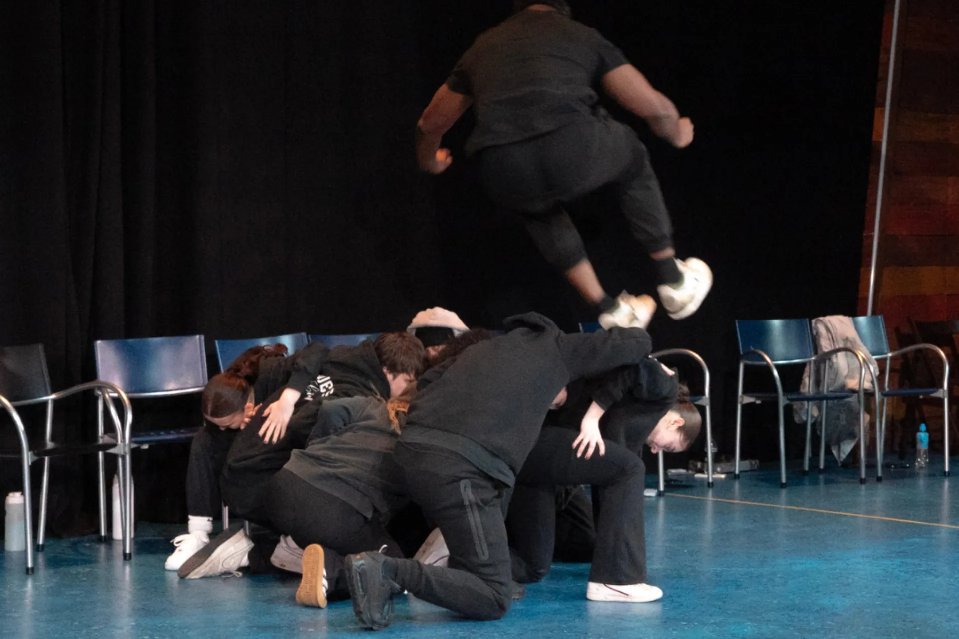 A group of dancers in black hoodies huddle together on the ground in a tight circle on a blue stage. Above them, another dancer is captured mid-air in a powerful jump. Blue chairs and a black backdrop frame the scene.