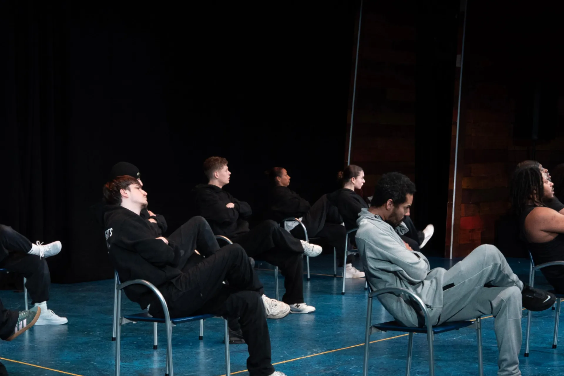 Dancers in black and grey streetwear sit separately in blue chairs across a blue stage. They are all leaning back with their arms crossed and legs folded, looking away in a synchronized, contemplative pose.