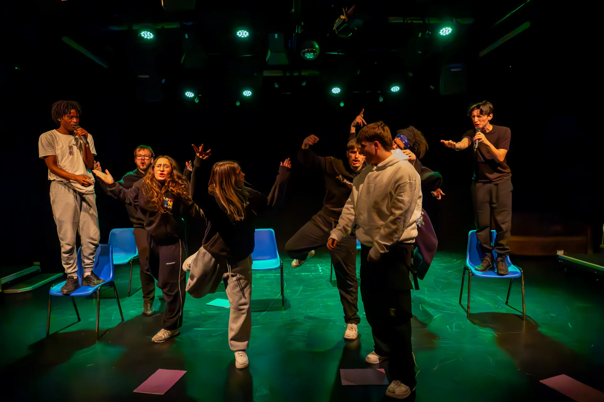 A group of young performers in casual streetwear act out a spirited scene under green stage lights, with two individuals standing on chairs and holding microphones.
