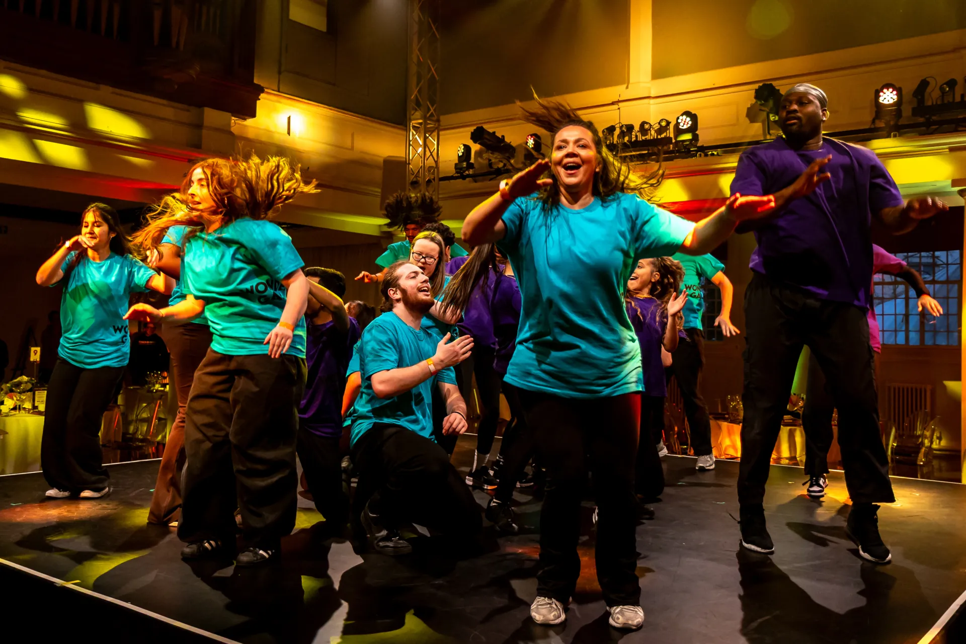 A joyful ensemble of performers in bright teal and purple t-shirts dance energetically on a stage during the "Wonder Gala," illuminated by warm yellow theatrical lighting.
